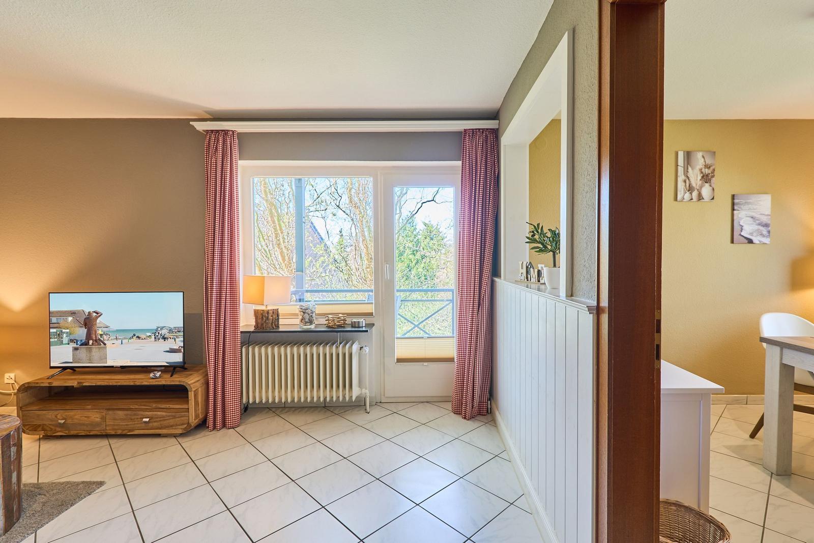 Wohnzimmer mit TV, Fenster und Balkon. Küche ist im Hintergrund sichtbar.