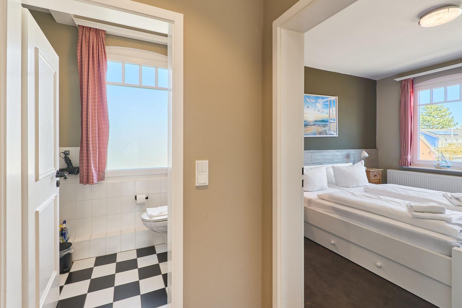 Schlafzimmer mit Bett und Fenster. Nebenraum mit Toilette und schwarz-weißem Boden.