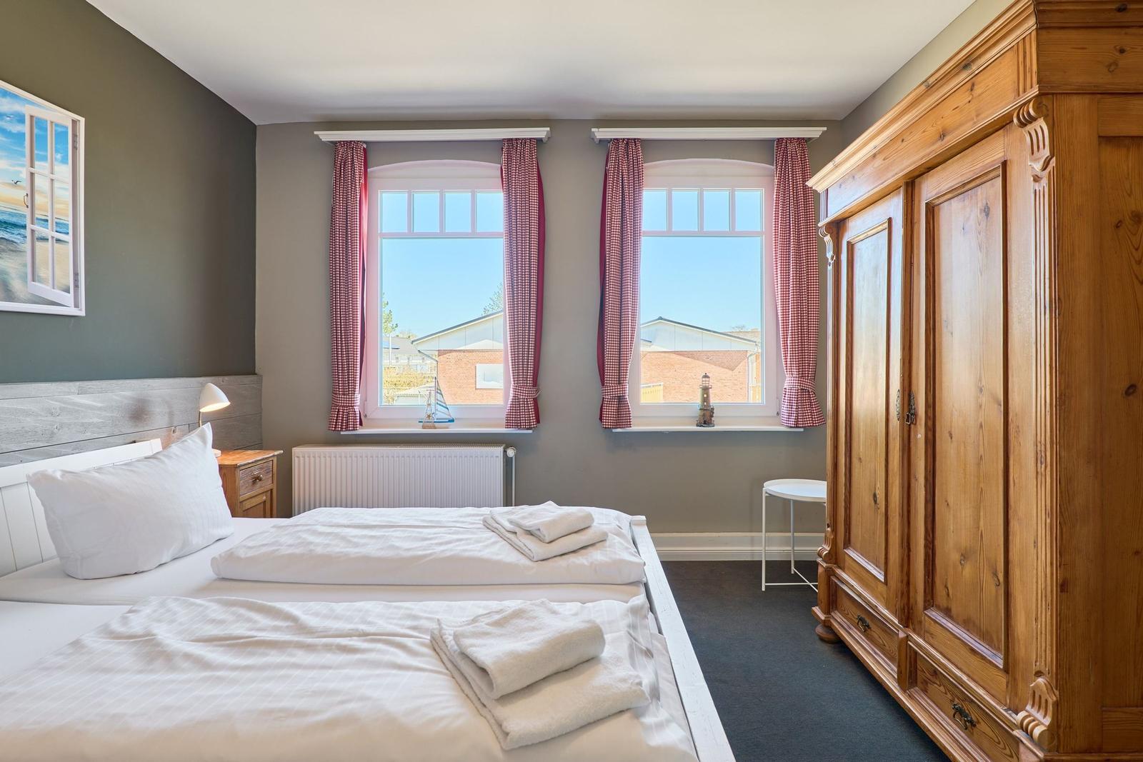 Zimmer mit Doppelbett, Holzschrank und zwei Fenstern mit roten Vorhängen.