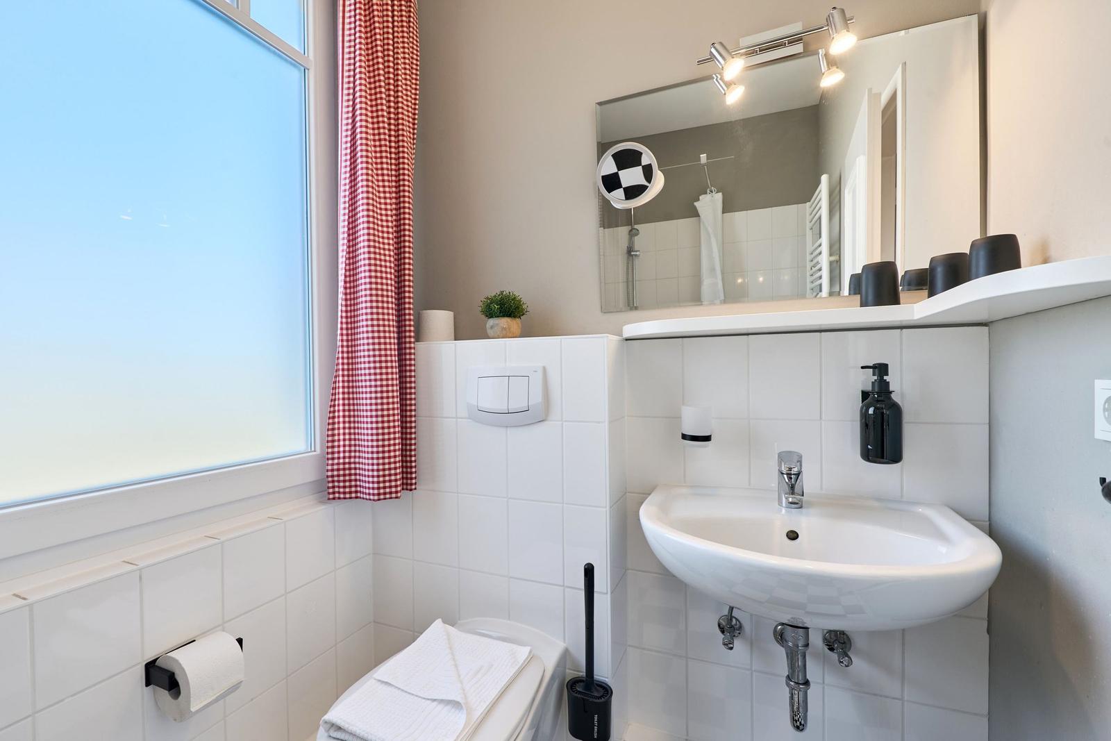 Badezimmer mit Waschbecken, Spiegel, Toilette und Fenster mit rotem Gittervorhang.