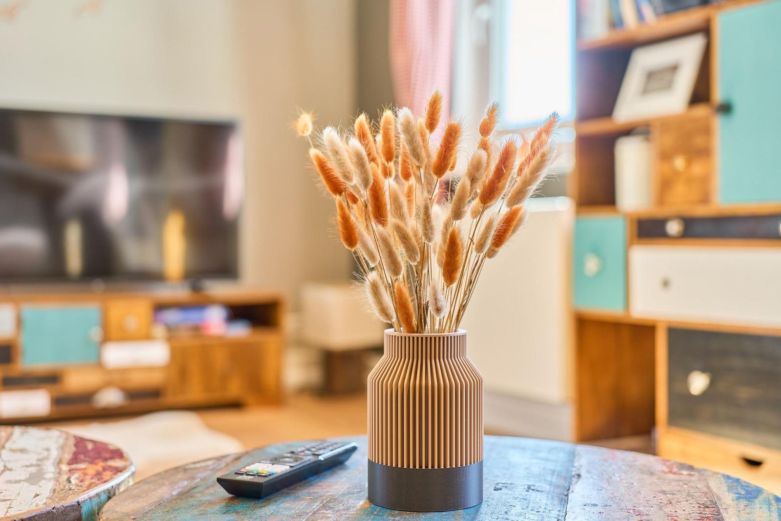 Ein Vase mit Trockenblumen steht auf einem Tisch. Im Hintergrund ist ein Fernseher und ein Regal zu sehen.
