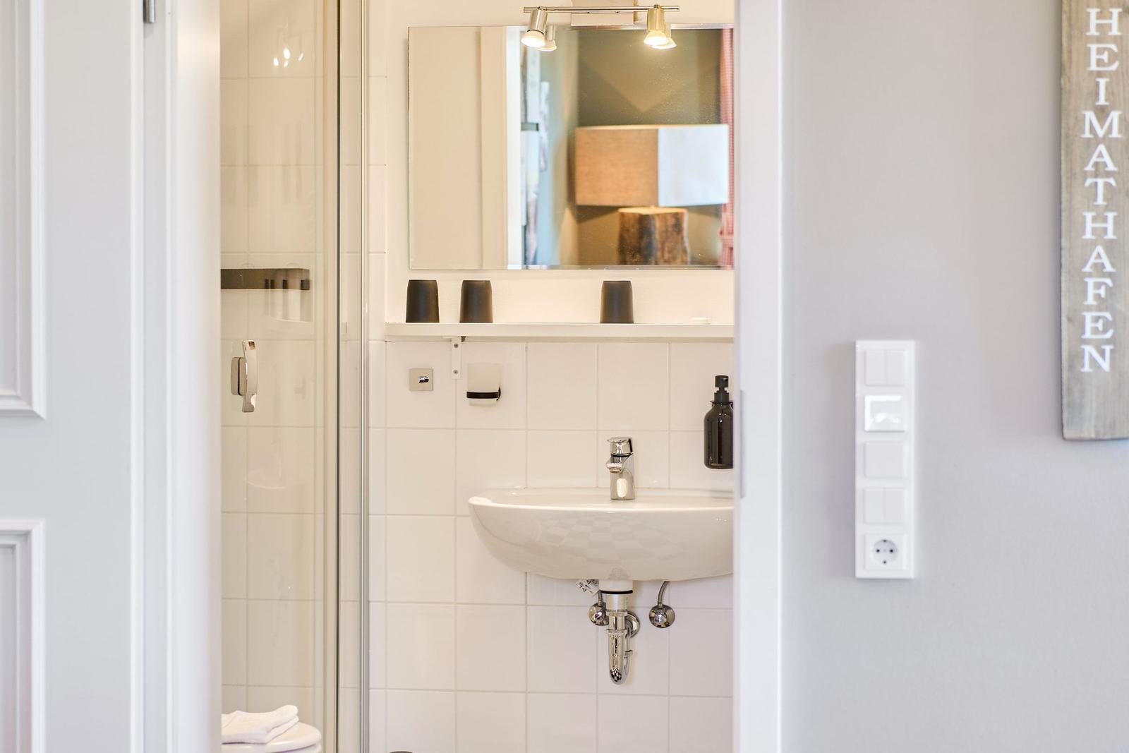 Badezimmer mit Dusche, Waschbecken und Spiegel. Wand mit Schalter und Schild.