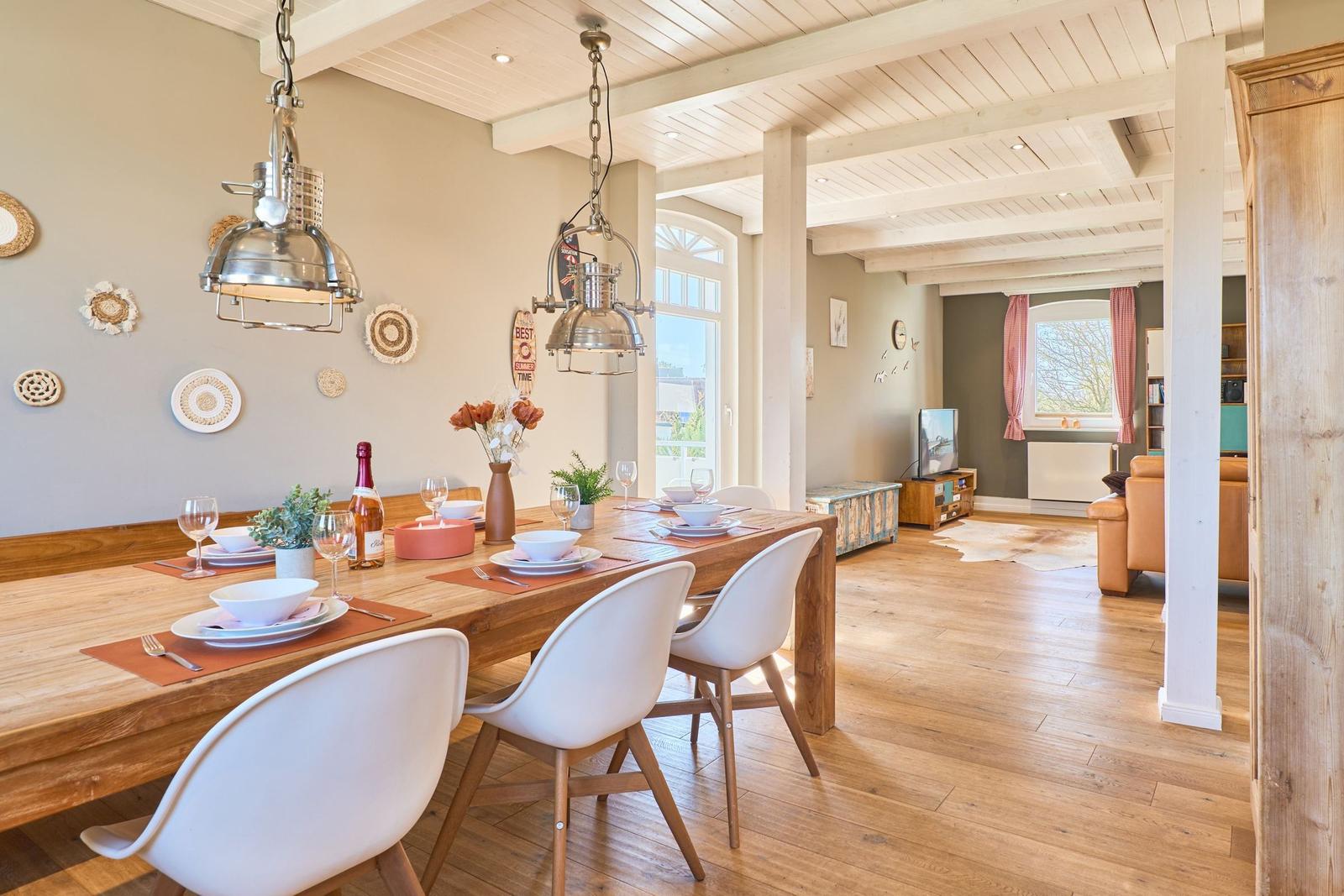 Essbereich mit Holztisch, weißen Stühlen und Deckenleuchten. Im Hintergrund Wohnzimmer mit Sofa und TV.