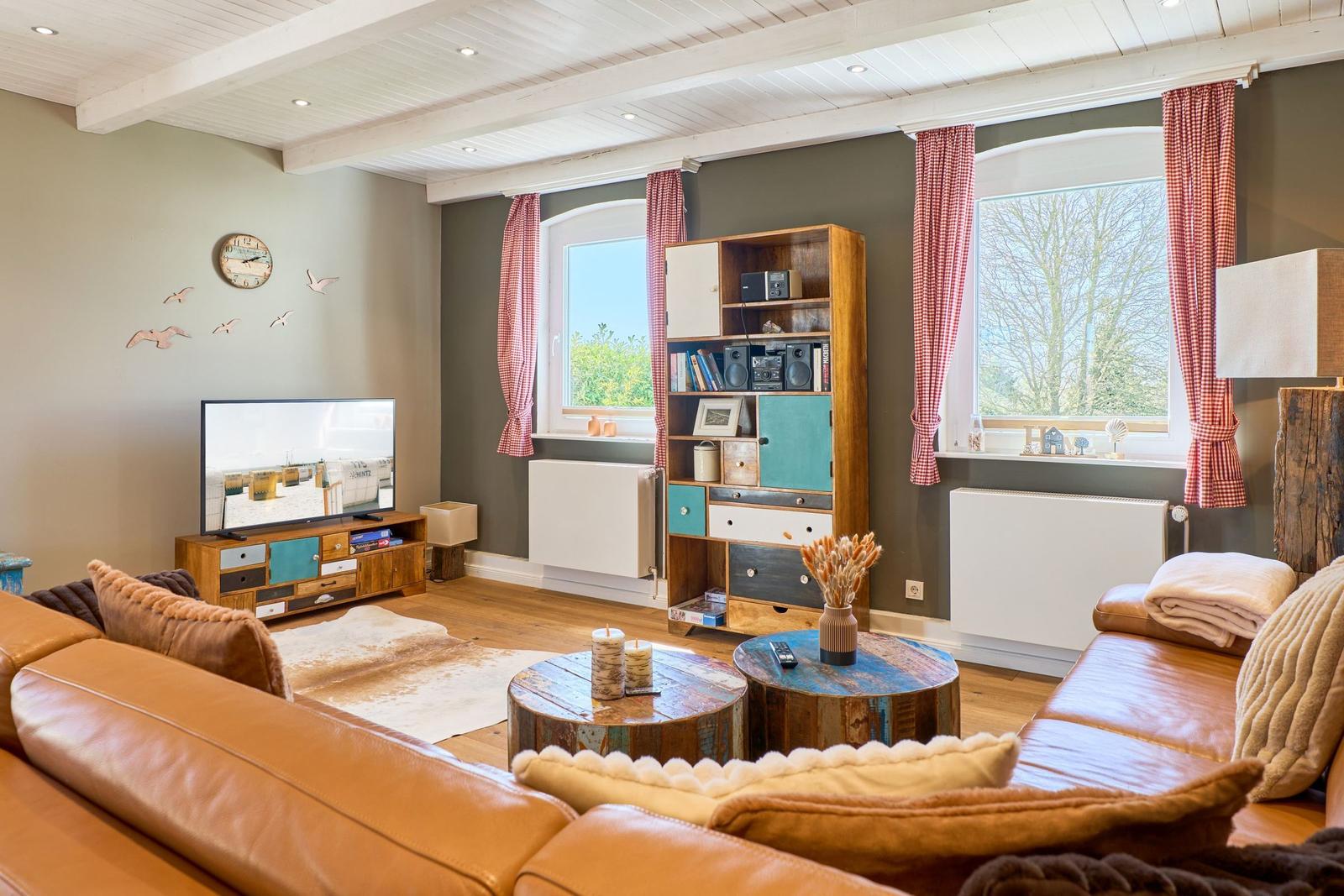 Ein Wohnzimmer mit braunem Ledersofa, Holztisch und Fernseher. An den Wänden hängen Vorhänge und ein Wanduhr.