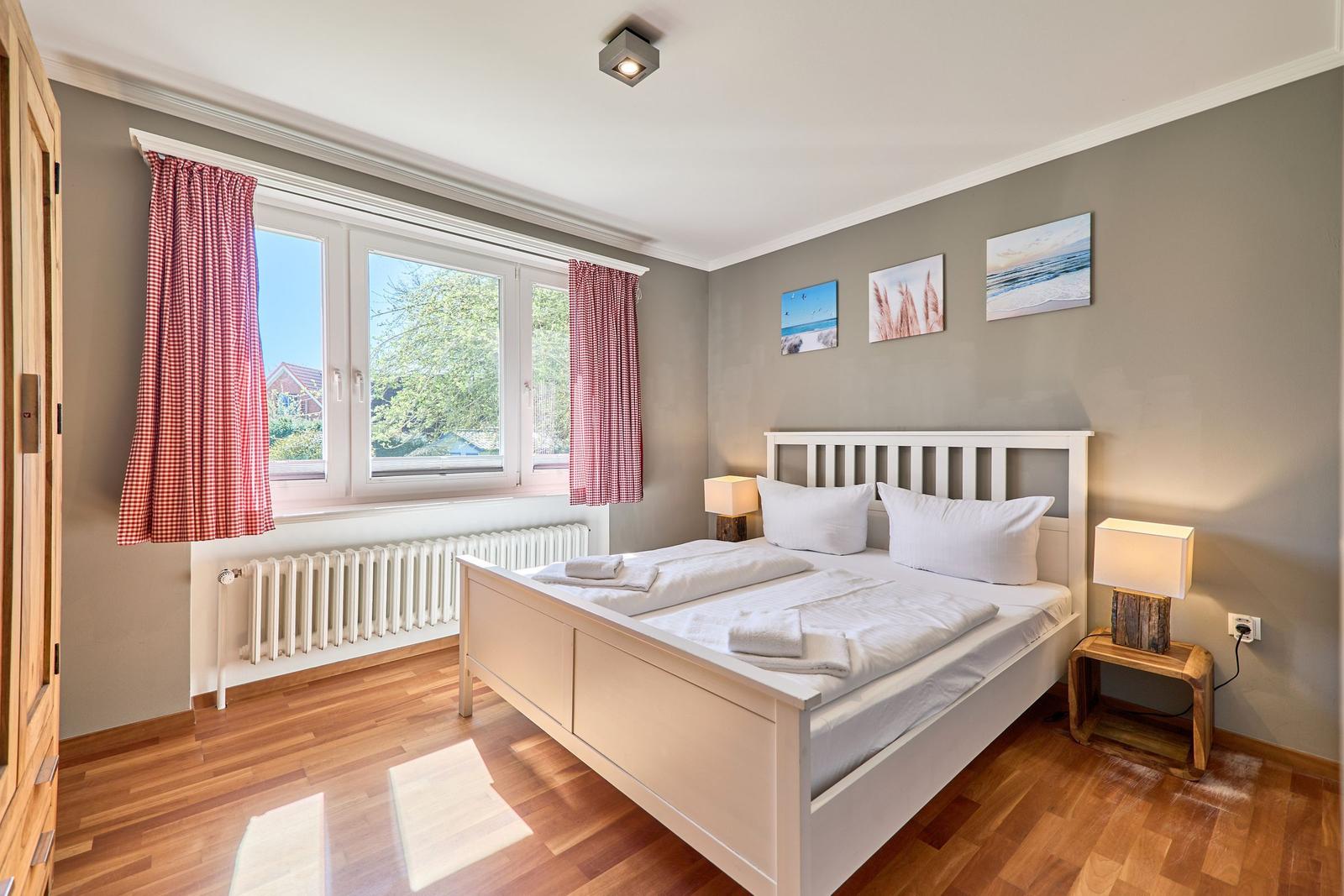 Schlafzimmer mit weißem Bett, Holzboden und Fenster mit roten Gardinen.