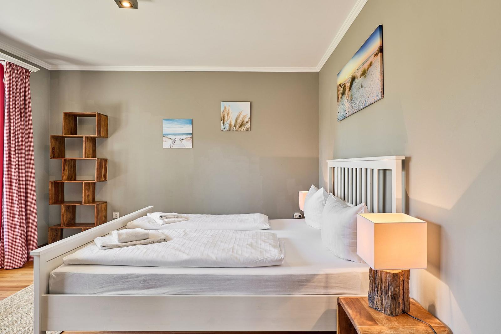 Schlafzimmer mit weißem Bett, Holzregal und Strandbildern an der Wand.