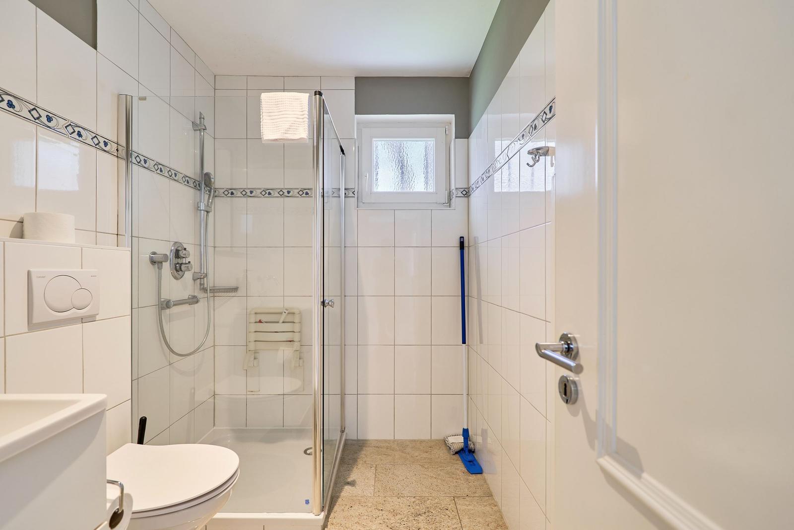 Badezimmer mit Dusche, Toilette und Waschbecken. Weiße Fliesen und kleine Fenster.