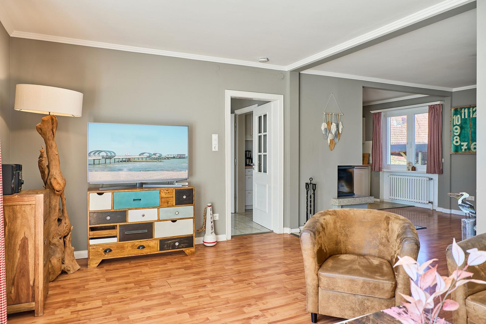 Wohnzimmer mit Fernseher, Sessel und Holzboden. Tür führt in Küche.