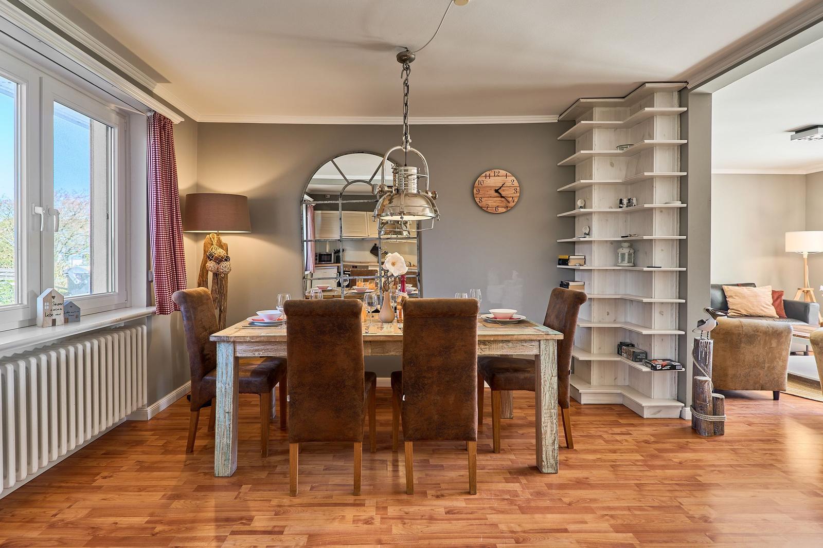 Esszimmer mit Holztisch, Stühlen und Bücherregal. Fenster mit Vorhängen und Wanduhr.