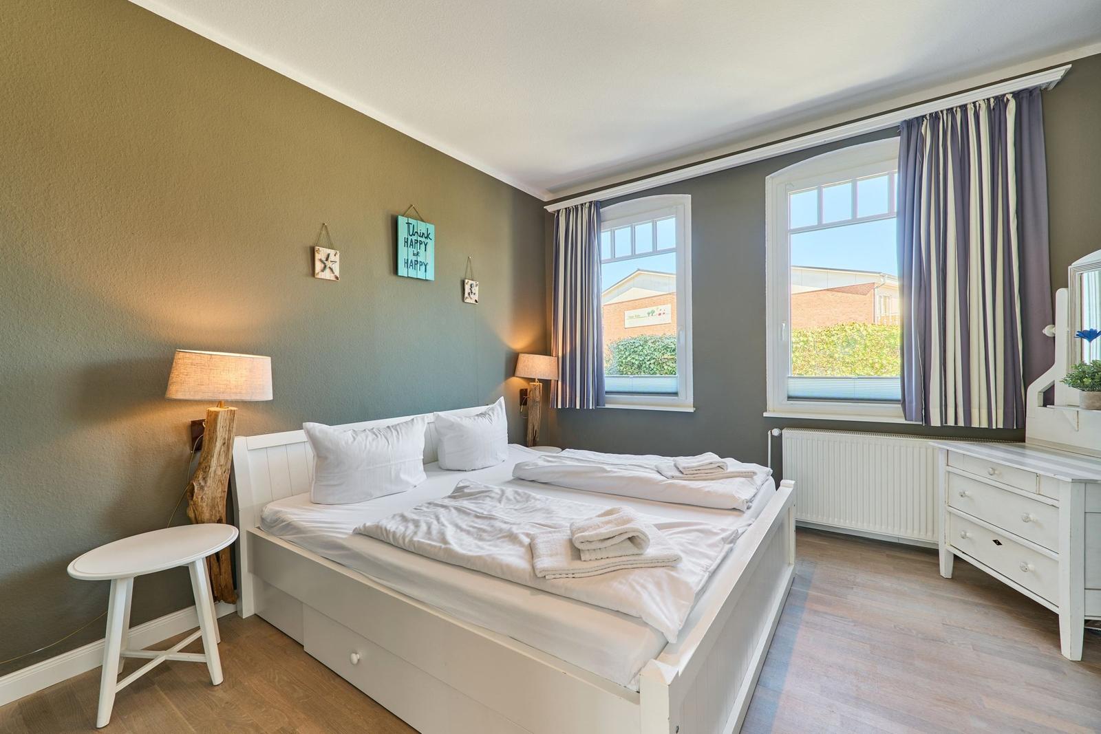 Schlafzimmer mit weißem Bett, Holzboden und zwei Fenstern mit Vorhängen.