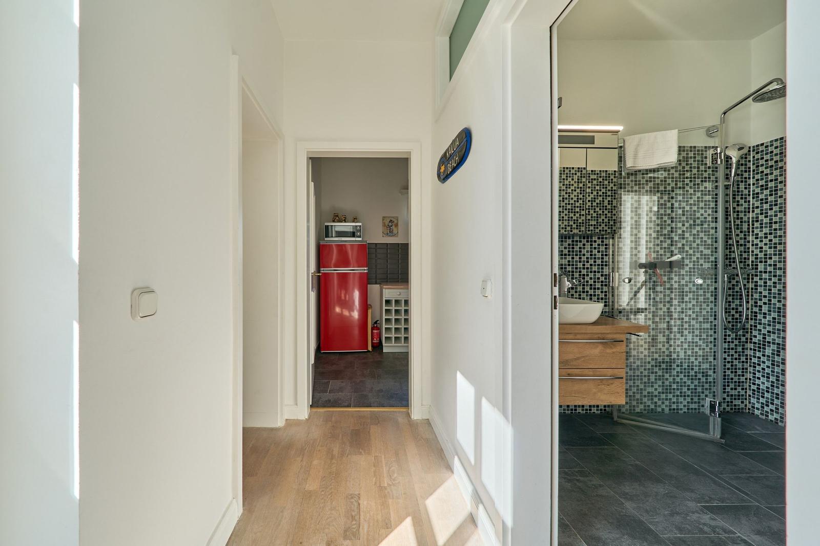 Flur mit Blick auf Küche und Bad. Küche hat roten Kühlschrank. Bad hat Dusche und Mosaikwand.
