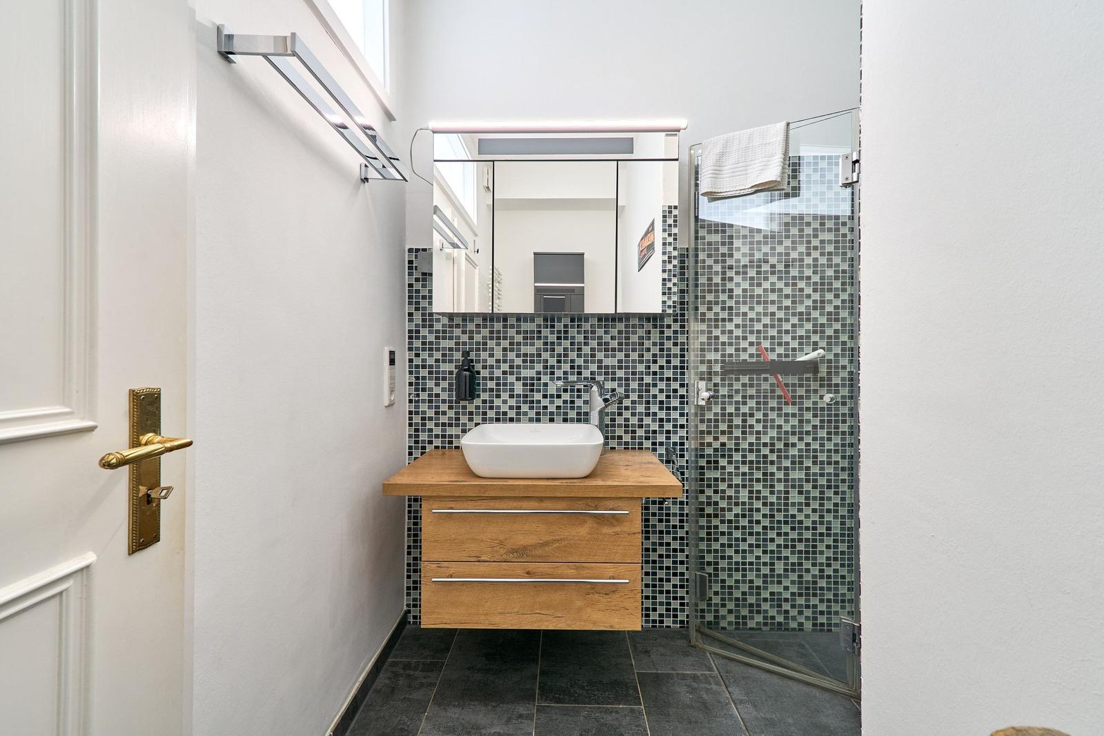 Badezimmer mit Waschbecken auf Holzkonsole, Spiegel und Dusche mit Mosaikwand.
