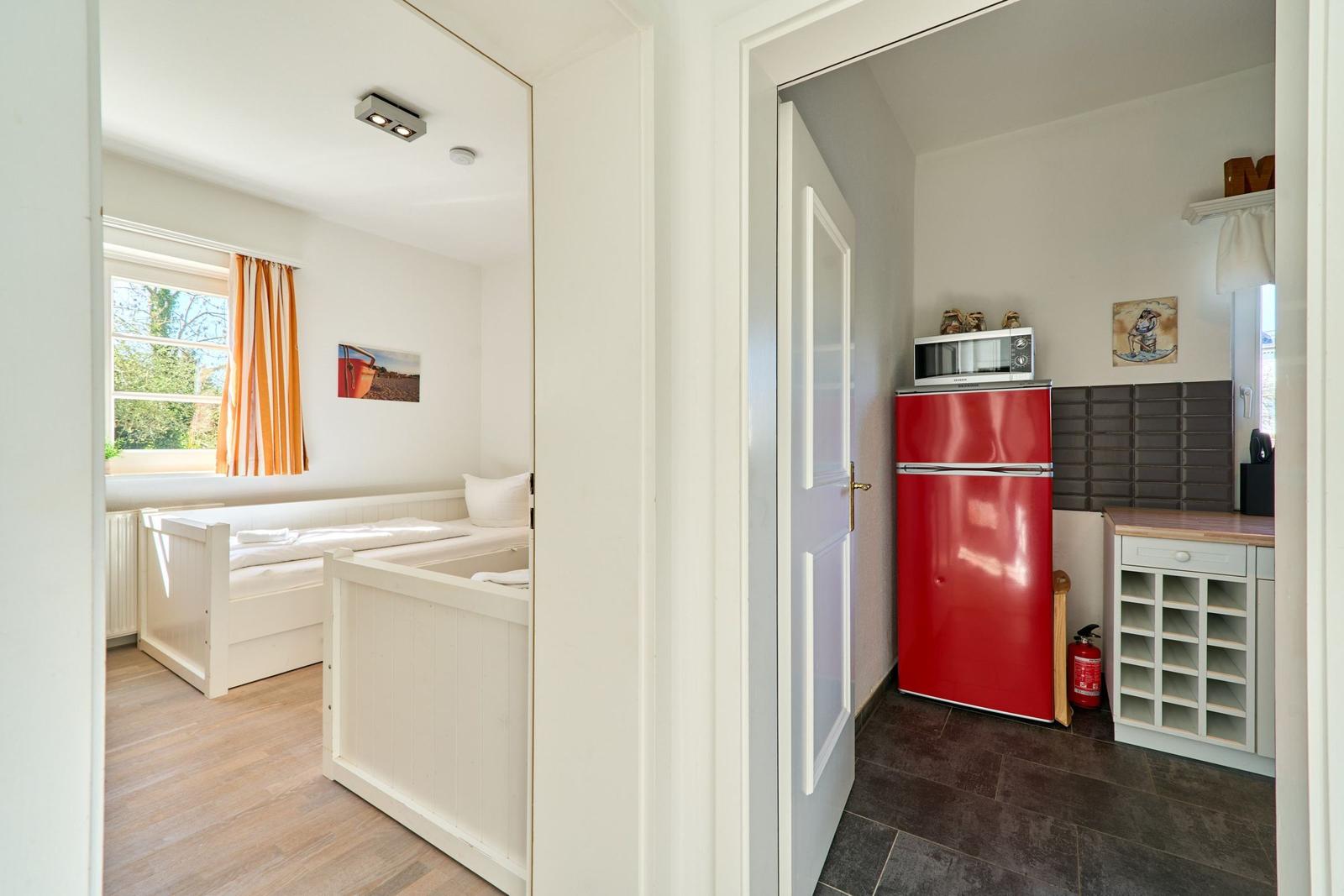 Schlafzimmer mit Bett und Fenster. Nebenraum mit rotem Kühlschrank und Mikrowelle.
