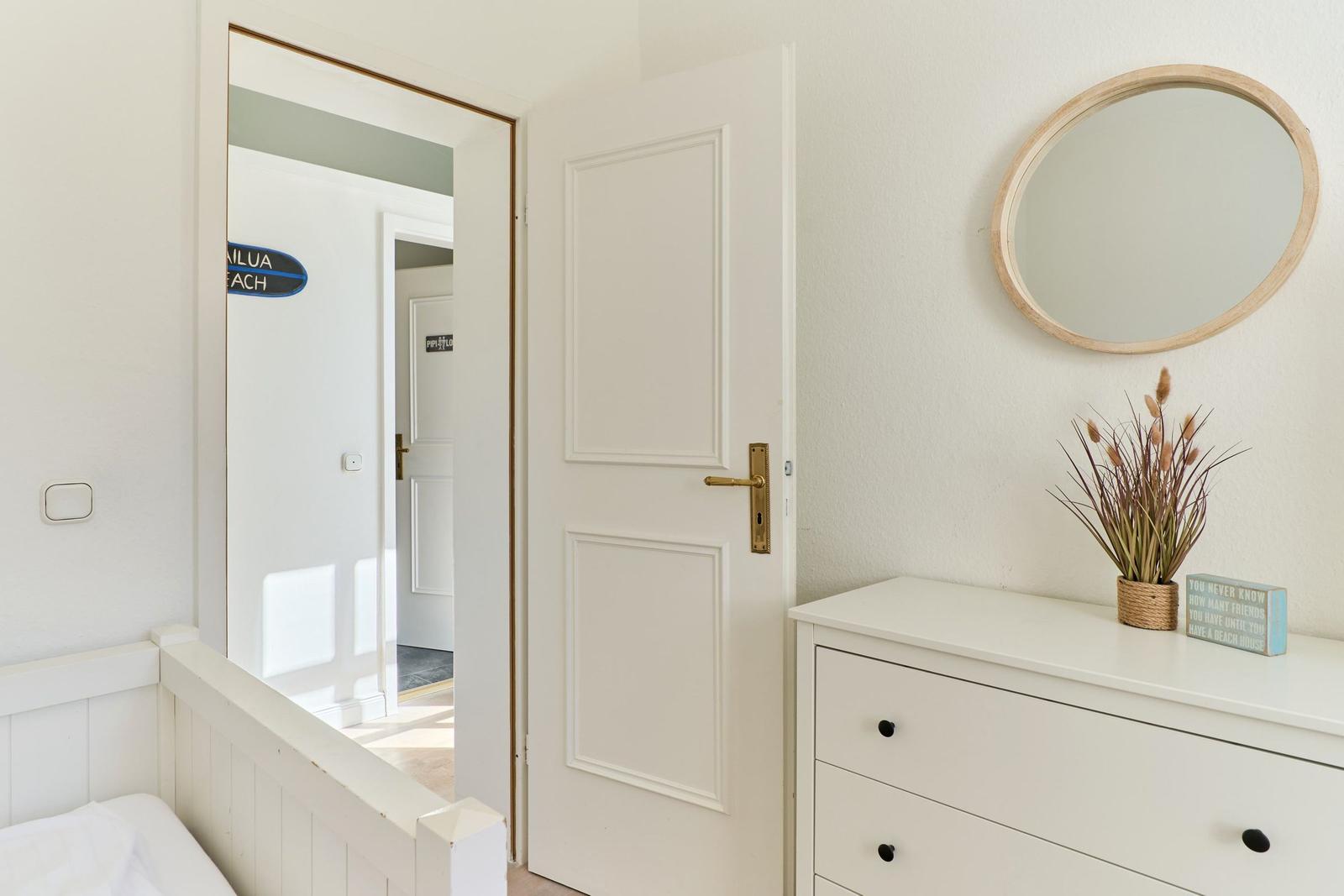 Schlafzimmer mit weißer Tür, Spiegelschrank und Pflanze. Blick in einen Flur mit Schild.