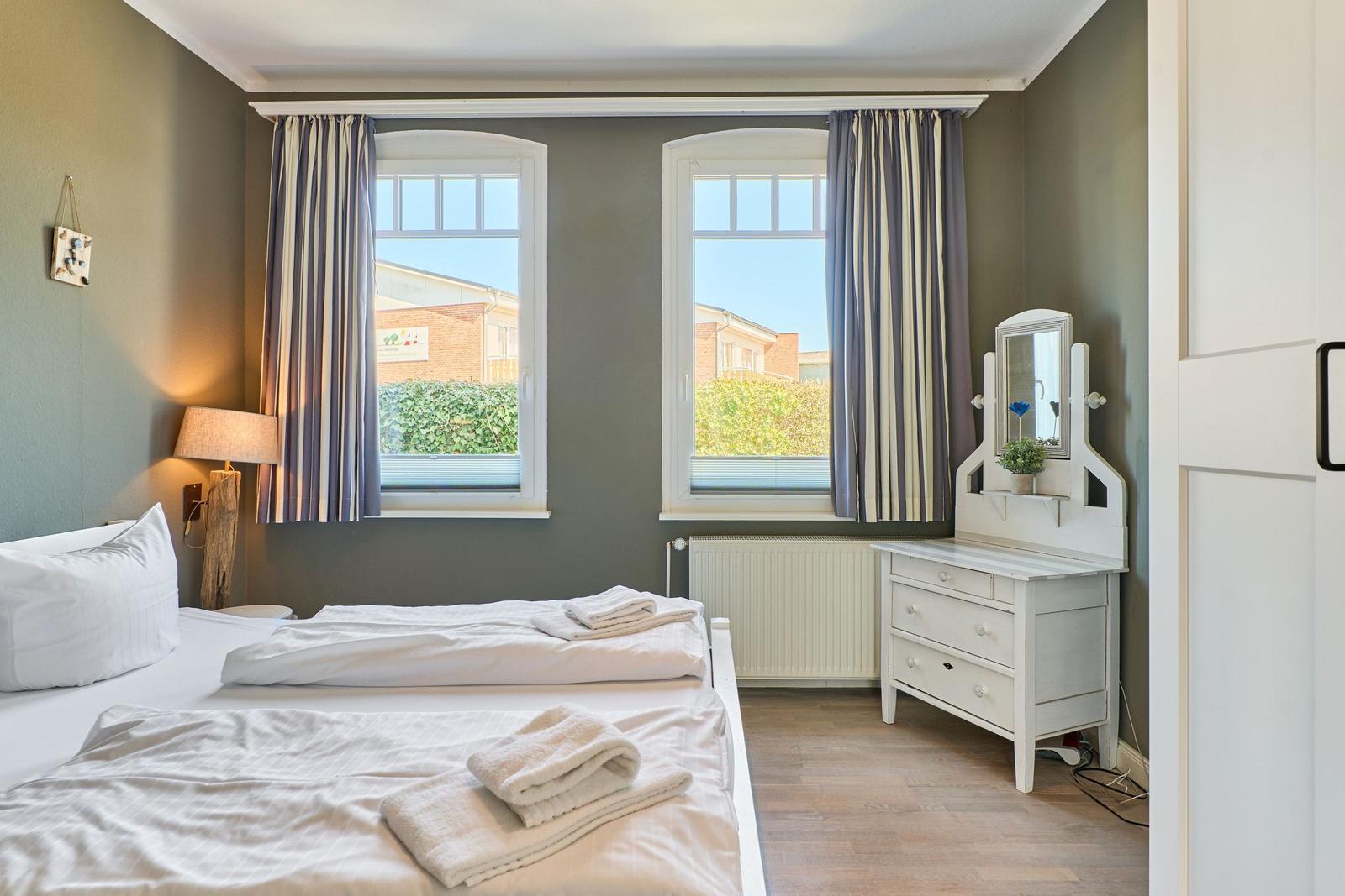 Zimmer mit Doppelbett, zwei Fenstern und Schrank mit Spiegel.