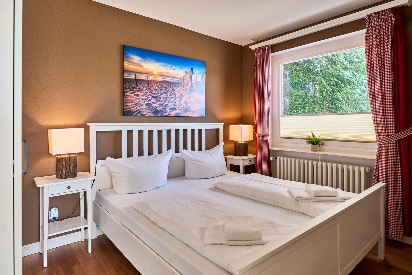 Schlafzimmer mit weißem Bett, zwei Nachttischen und Fenster mit roten Gardinen.