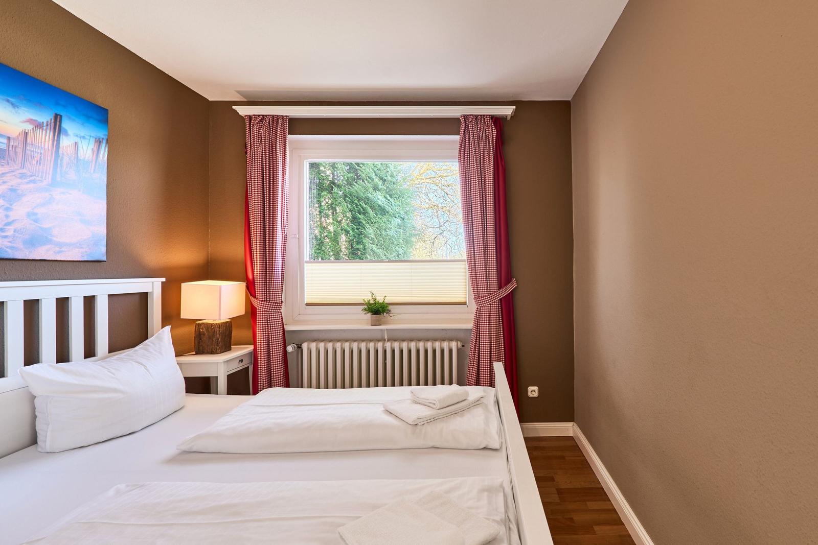 Ein Schlafzimmer mit weißem Bett, Fenster mit roten Vorhängen und Wandbild.