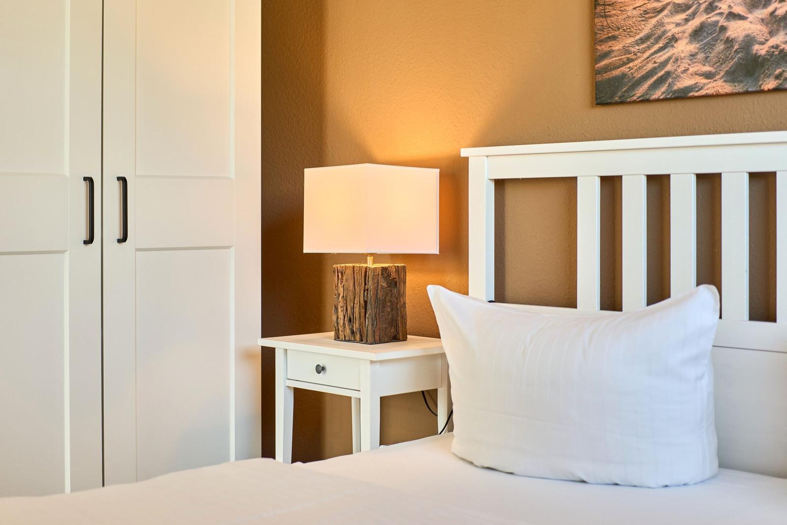 Schlafzimmer mit weißem Bett, Nachttisch und Lampe auf Holzsockel.