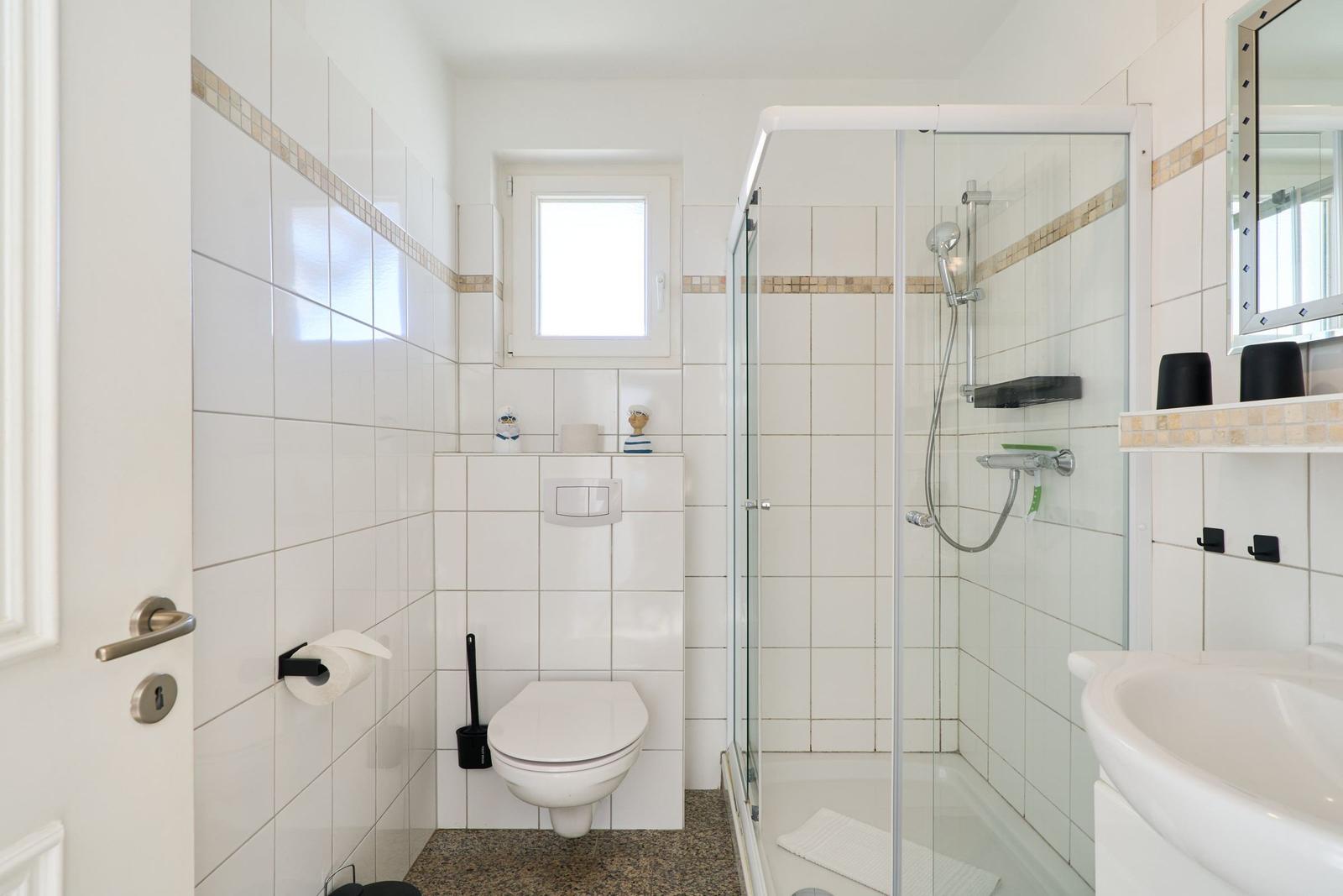 Badezimmer mit Dusche, Toilette und Waschbecken. Weiße Fliesen und kleine Fenster.