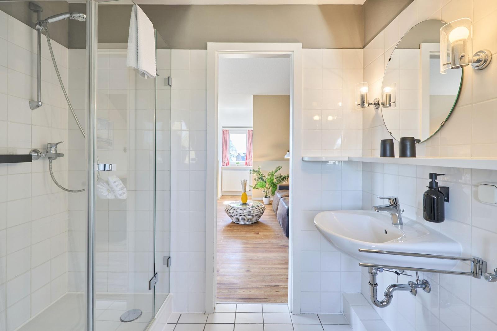 Badezimmer mit Dusche, Waschbecken und Blick in das Wohnzimmer.