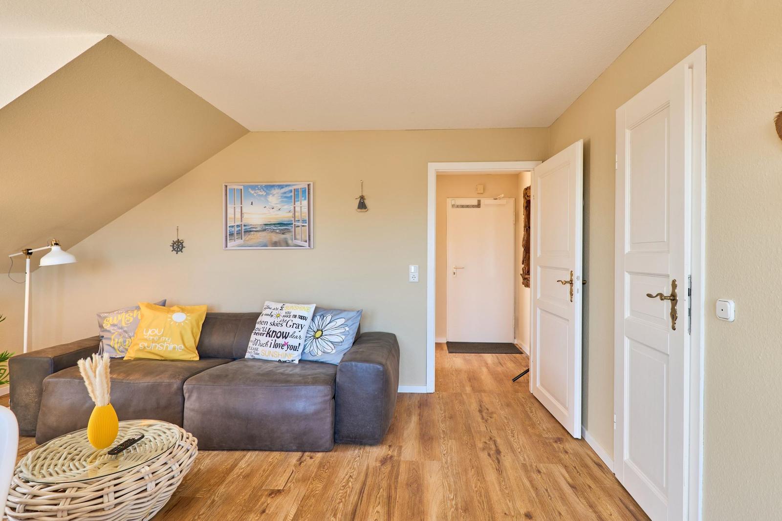 Wohnzimmer mit Sofa, Kissen, Wandbild und Holzboden. Tür führt in den Flur.
