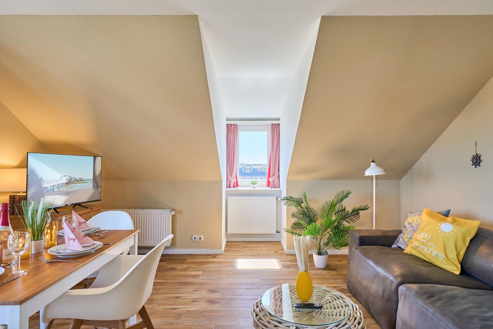 Wohn- und Essbereich mit Sofa, Tisch und Fenster. Holzboden und schräge Decke.