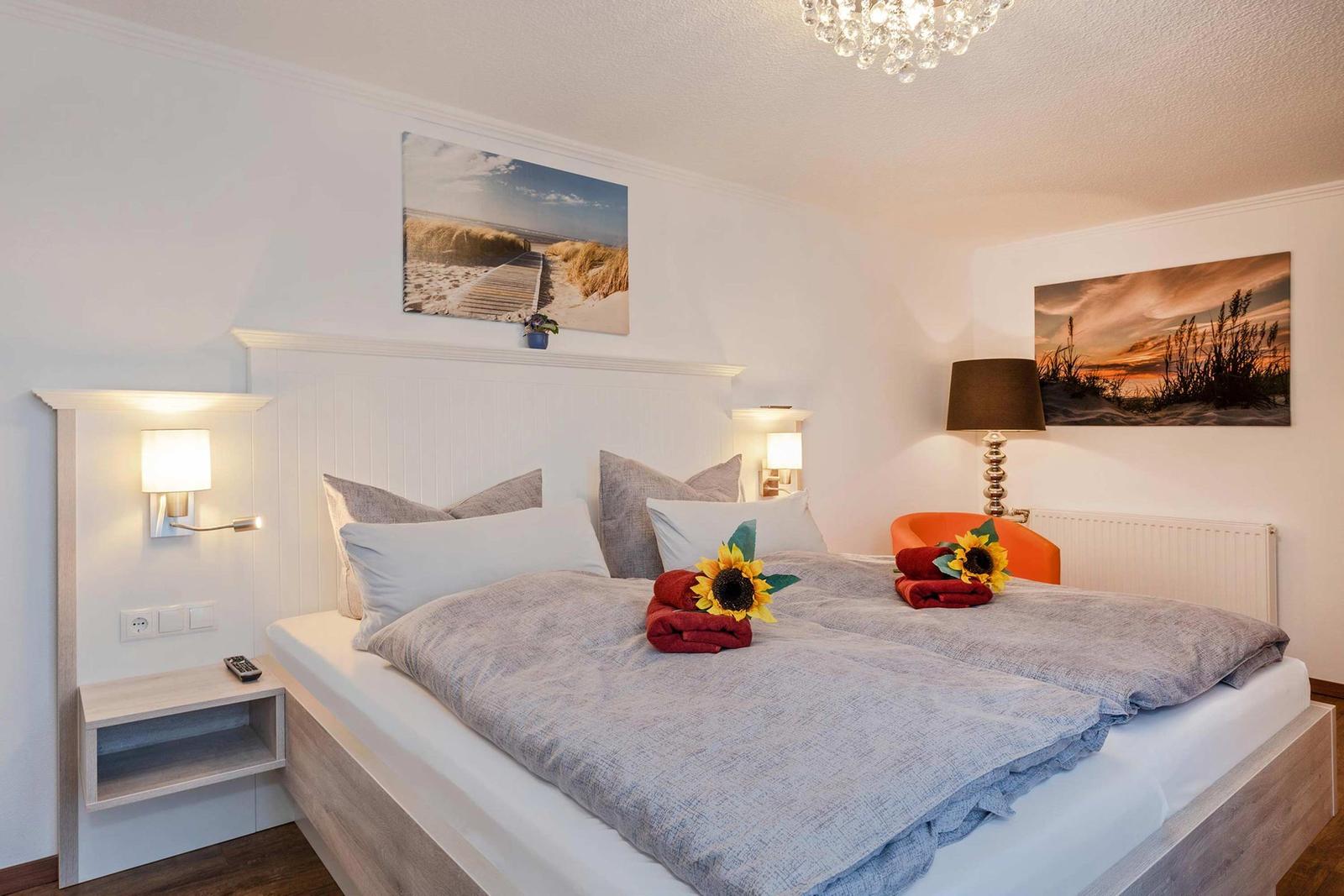 Schlafzimmer mit Bett, Wandbildern und Nachttisch. Auf dem Bett liegen Handtücher mit Sonnenblumen.