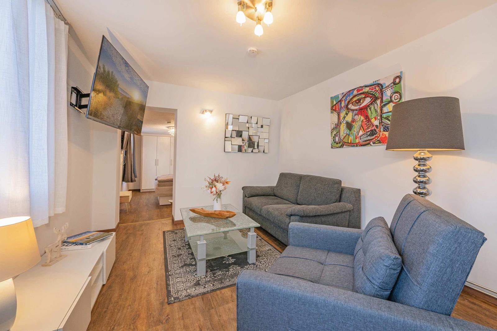 Wohnzimmer mit Sofa, Sessel, Glas-Tisch und Wandbildern.