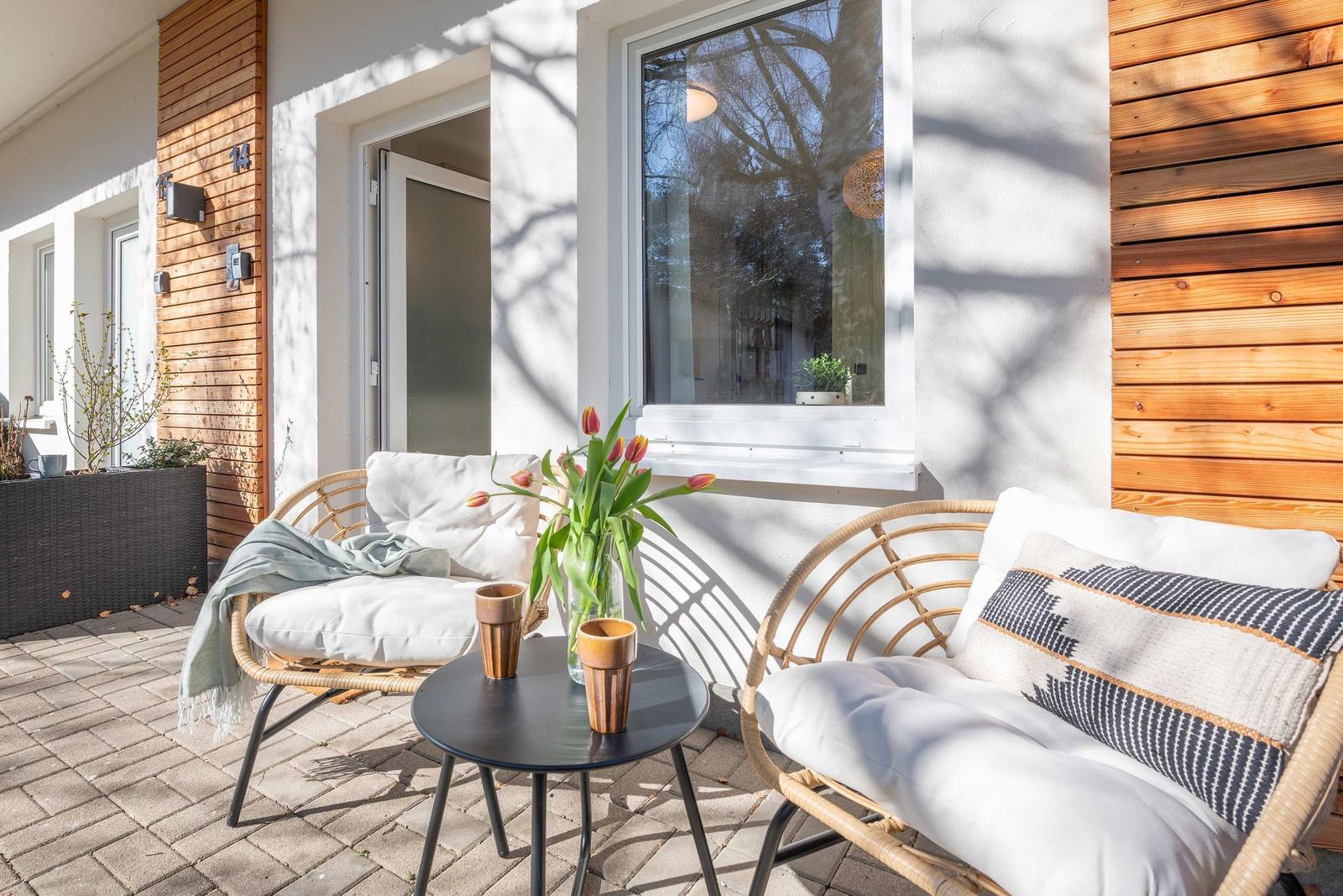 Terrasse mit zwei Sesseln, Tisch und Blumen vor einem Fenster.