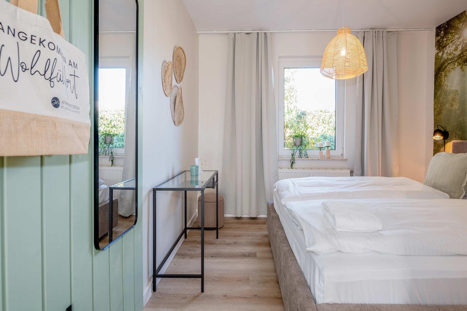 Schlafzimmer mit Bett, Fenster, Lampe und Spiegel an der Wand.