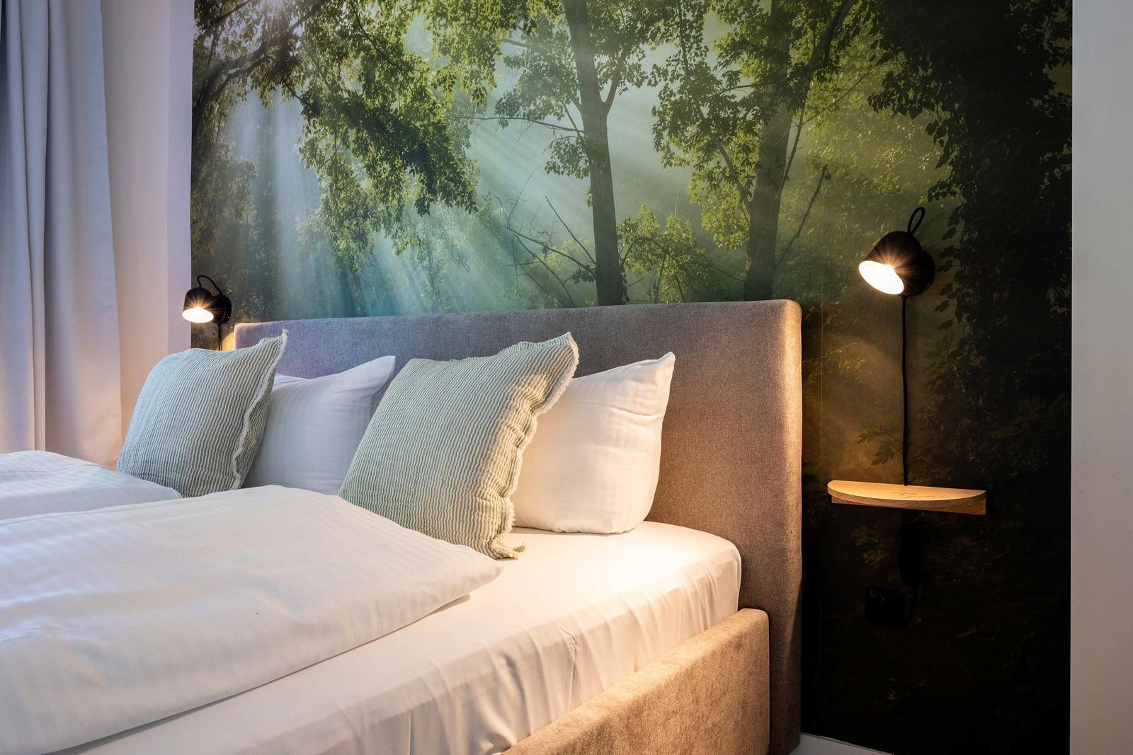 Schlafzimmer mit Bett, Kissen und Wandbild von Wald. Beleuchtung an beiden Seiten.