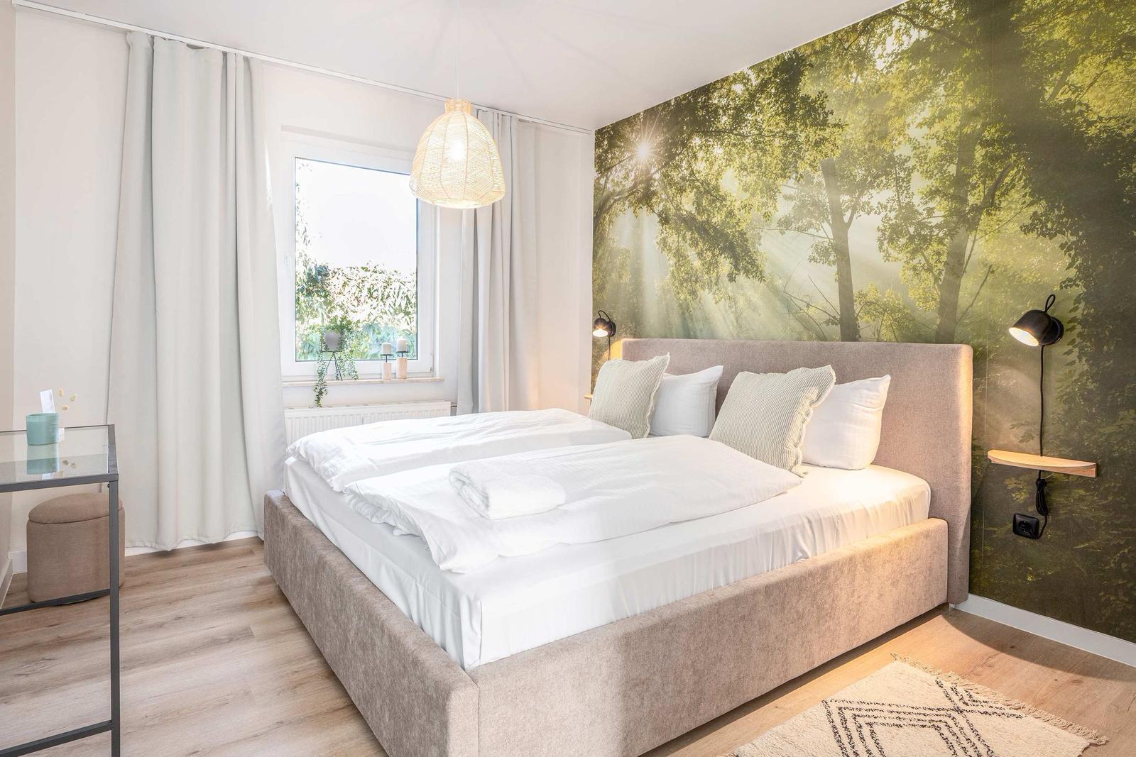 Schlafzimmer mit Bett, Fenster und Wandbild mit Waldmotiv.