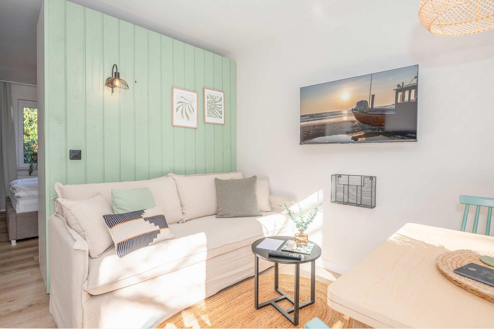 Wohnzimmer mit Sofa, Tisch und Wandbild. Grüne Holzwand und Essbereich sichtbar.