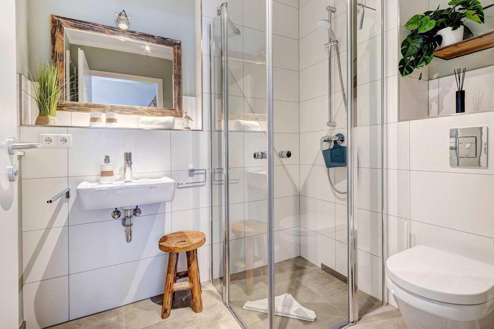 Badezimmer mit Dusche, Waschbecken und Toilette. Holzstuhl und Pflanzen sichtbar.