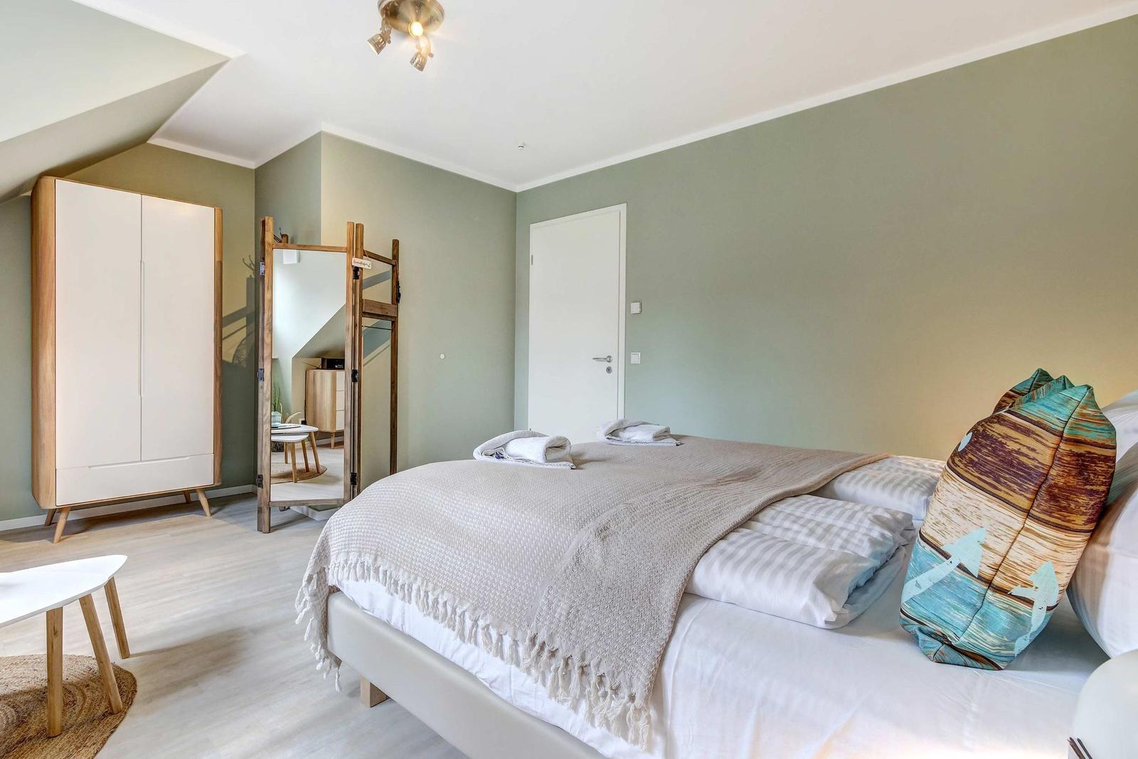 Schlafzimmer mit Bett, Kleiderschrank und Spiegel. Wandfarbe grün, Boden hell.