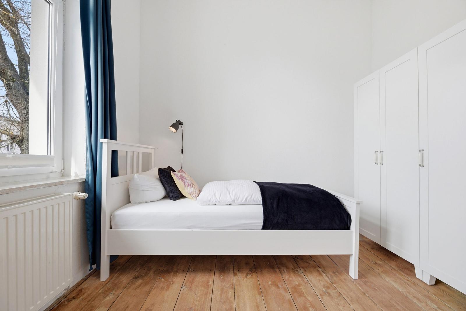 A bedroom with a white bed, blue curtains, and wooden floor. A white wardrobe is on the wall.