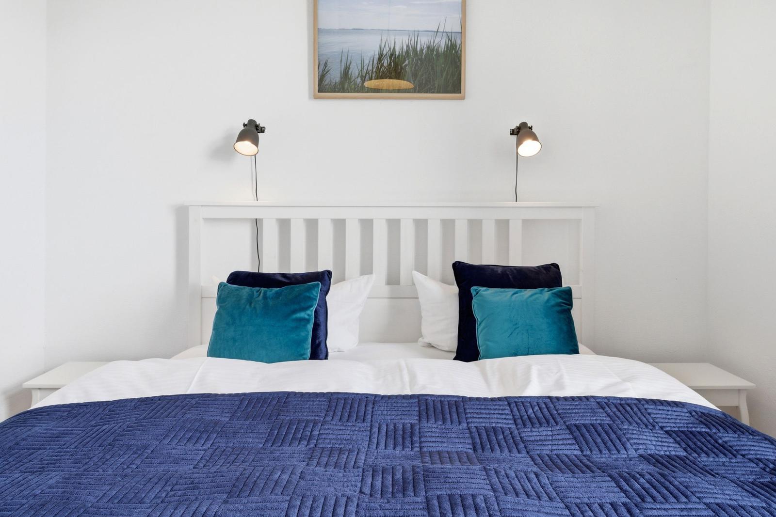 A bed with blue quilt and pillows, white wall, two wall lamps, and a picture above the headboard.
