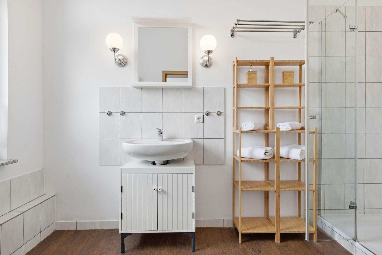 Bathroom with sink, mirror, shower, and wooden shelf with towels.