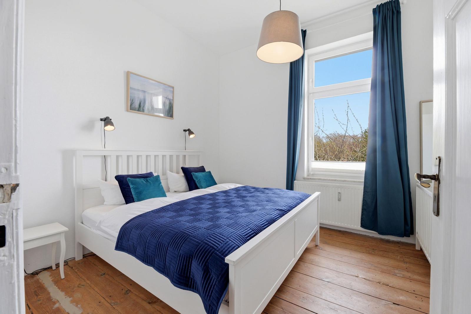 Bedroom with white bed, blue quilt, and wooden floor. Window with blue curtains.