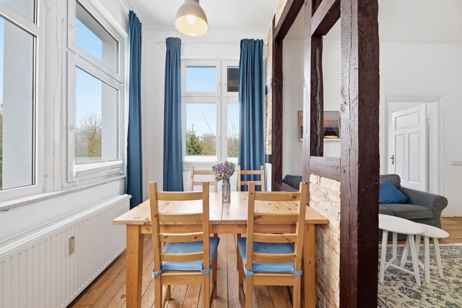 Dining area with wooden table, blue chairs, and windows. Living area with sofa in background.
