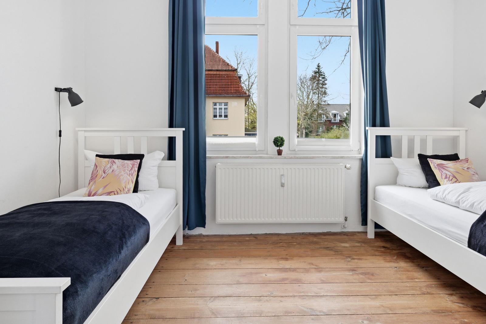 Two single beds with white linens and dark blankets stand before a window with blue curtains.