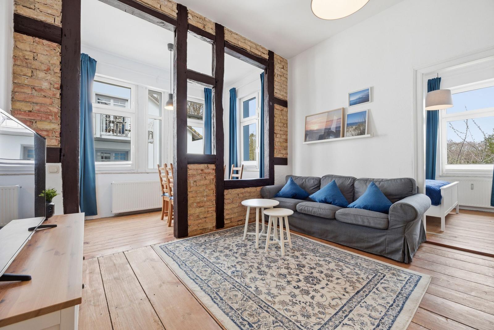 Living room with sofa, rug, and wooden floor. Passage to dining area with wooden chairs.