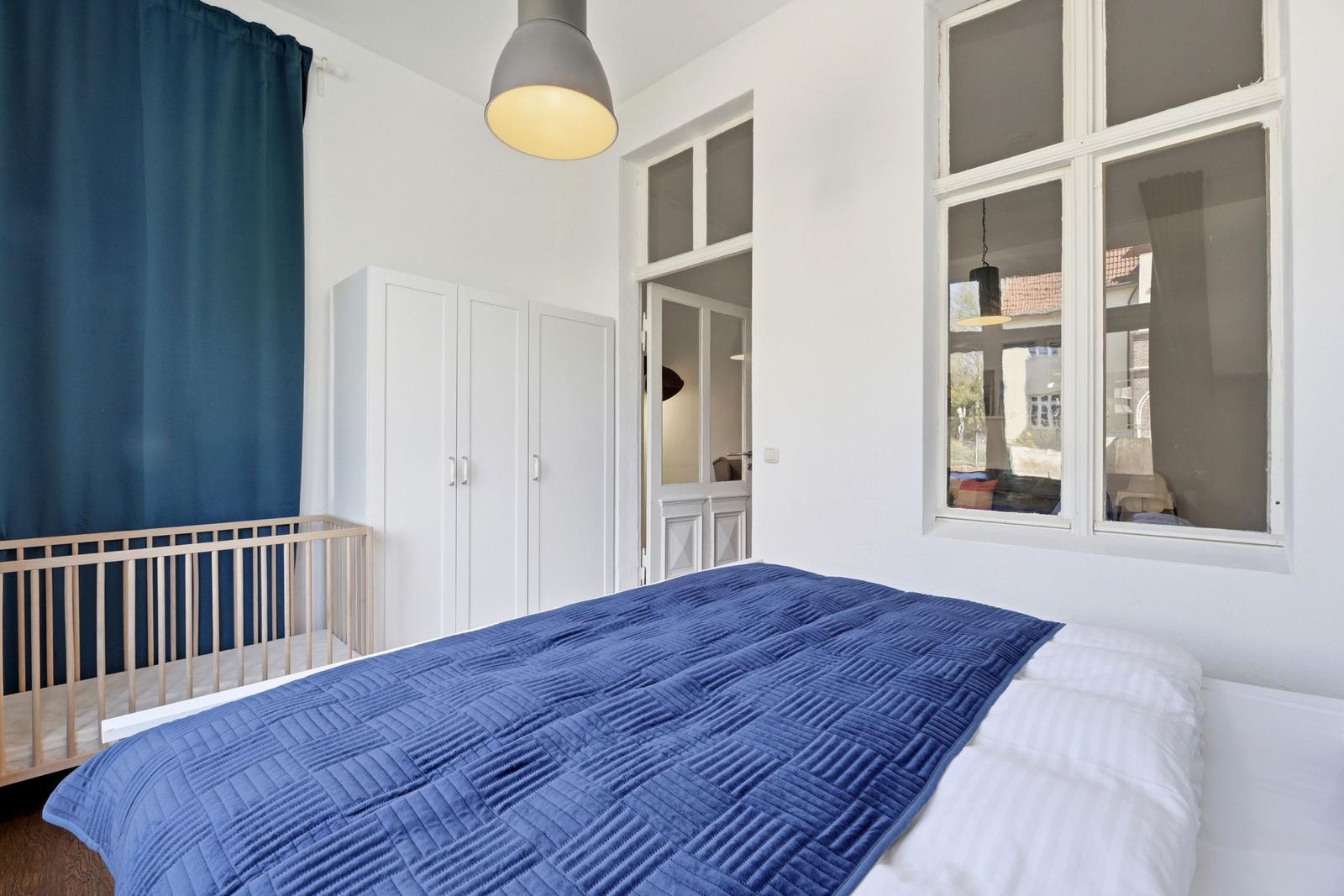 Bedroom with bed, blue cover, white walls, and windows. A wardrobe is against the wall.