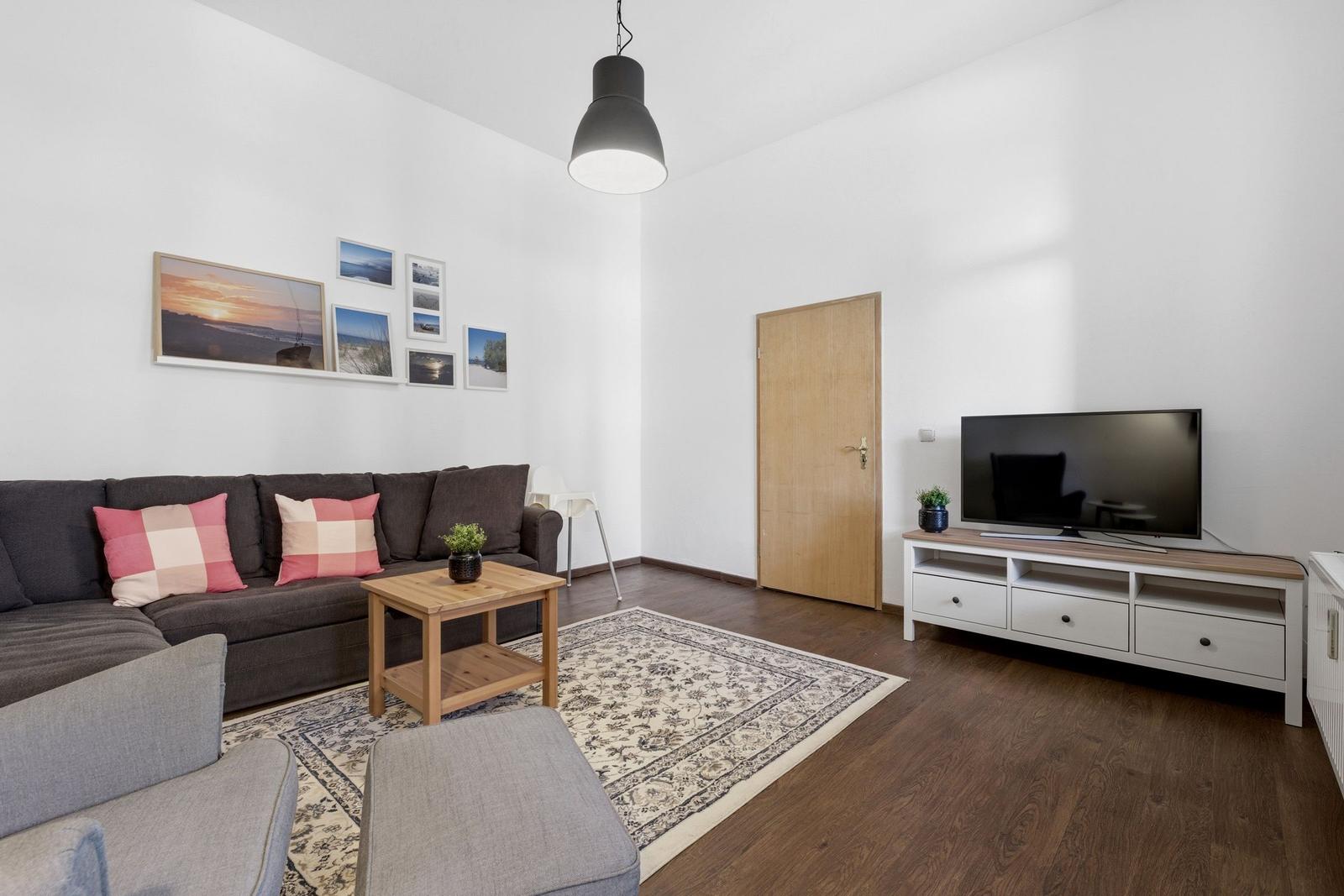 Living room with sofa, TV, and wooden floor. Wall art and rug visible.
