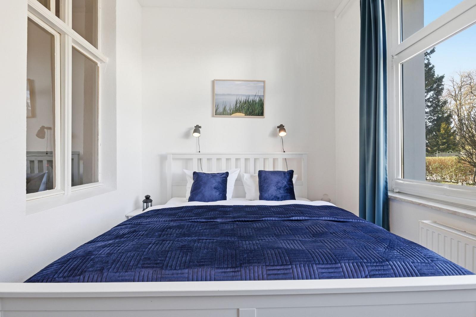 Bedroom with white bed, blue quilt, and two windows.