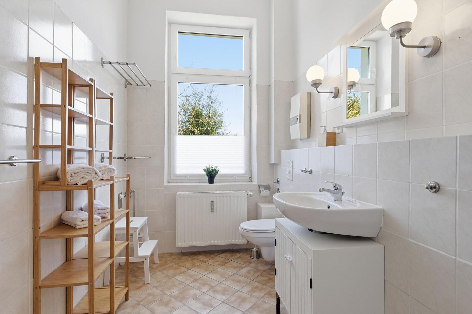 Badezimmer mit Waschbecken, Toilette, Fenster und Holzregal für Handtücher.