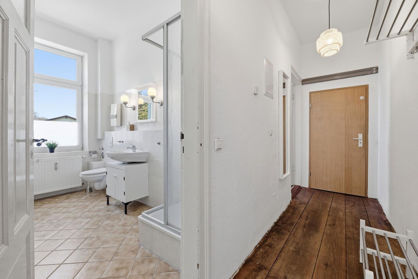 Badezimmer mit Dusche, Waschbecken und Toilette. Flur mit Holzboden und Tür.