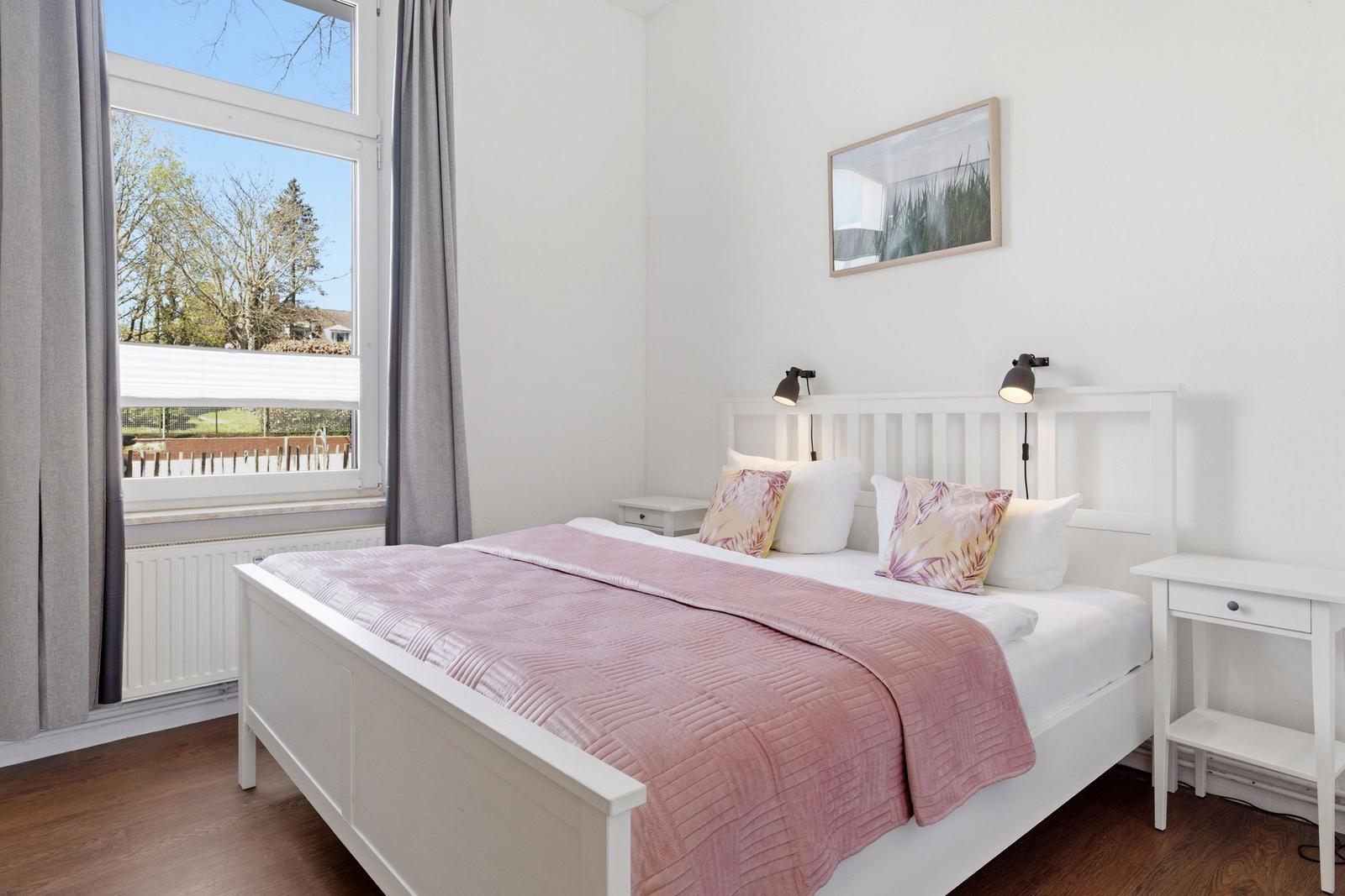 Schlafzimmer mit weißem Bett, rosa Decke und Blick auf Garten.