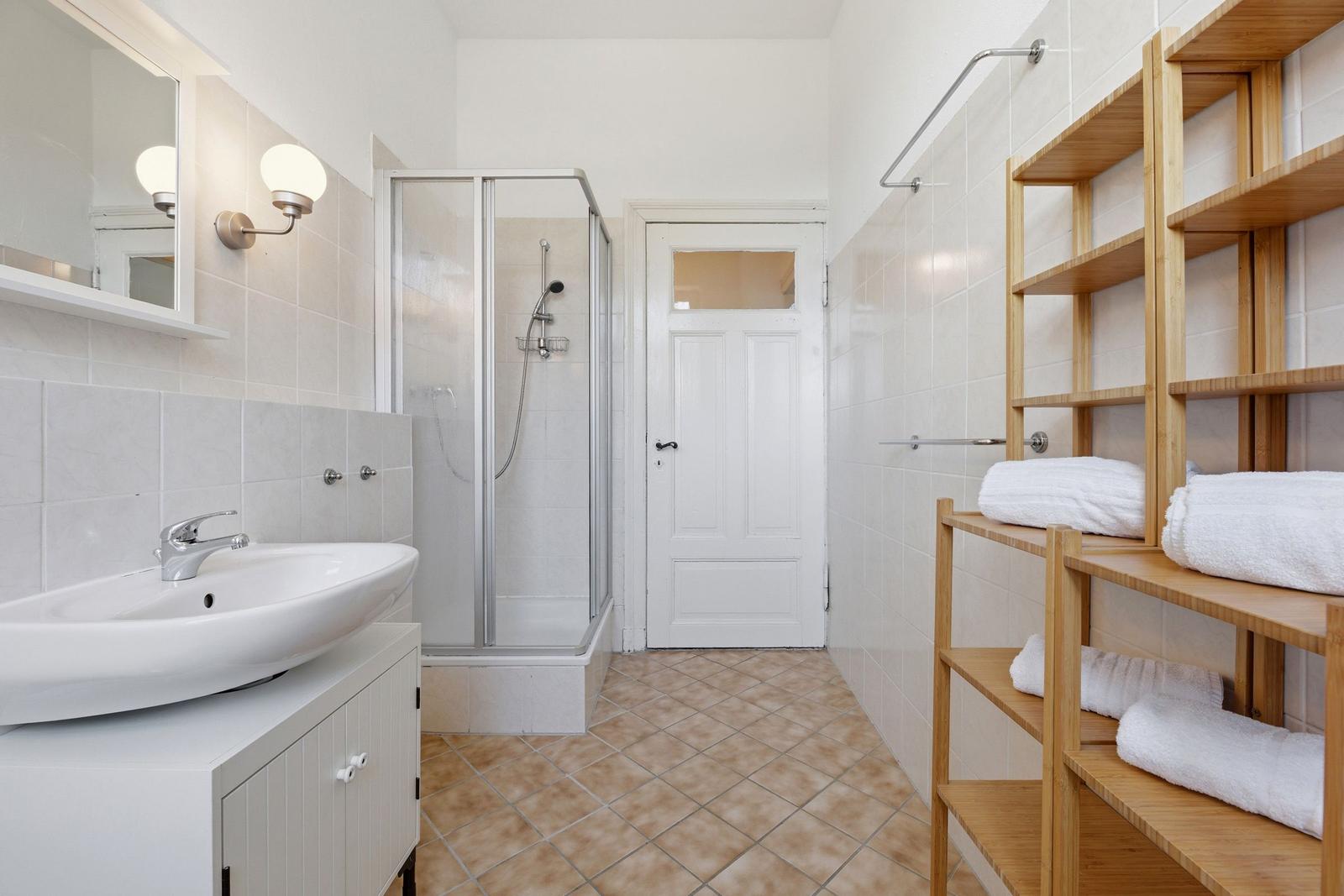 Badezimmer mit Dusche, Waschbecken und Holzregal mit Handtüchern.