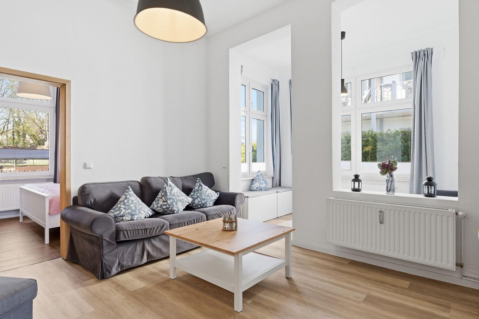 Wohnzimmer mit Sofa, Couchtisch und Fenstern. Blick in Schlafzimmer.